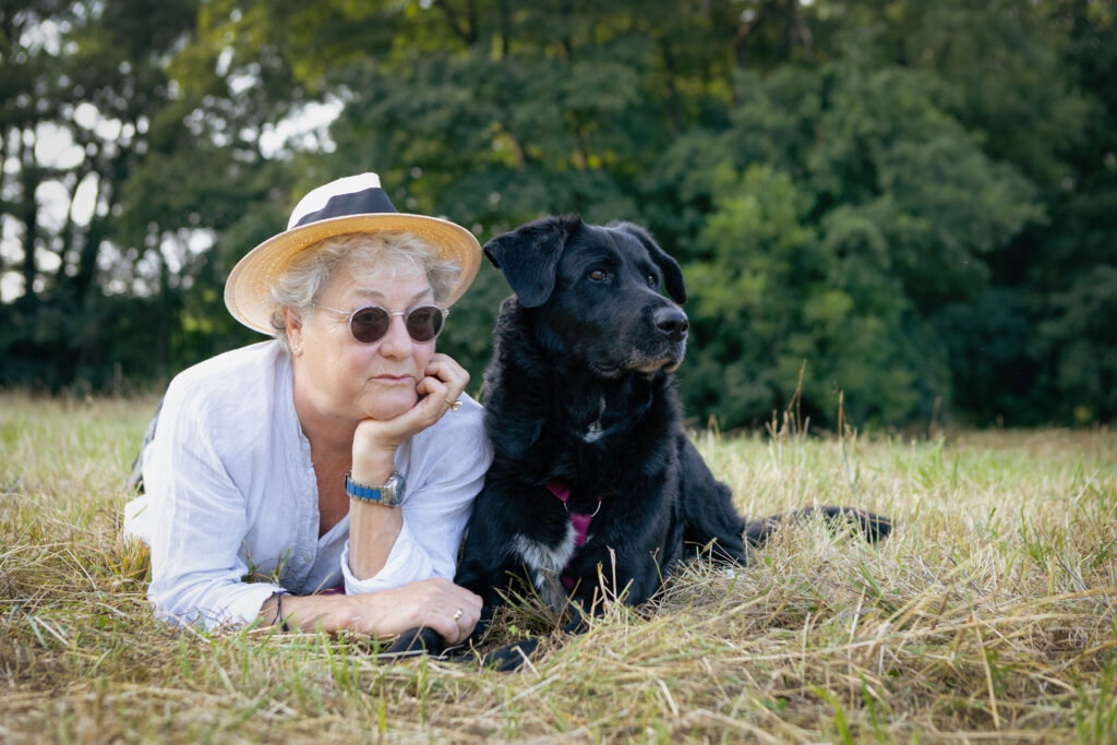 Besitzerin in weiß gekleidet liegt mit ihrem schwarzen Hund im Gras und beide blicken nach rechts