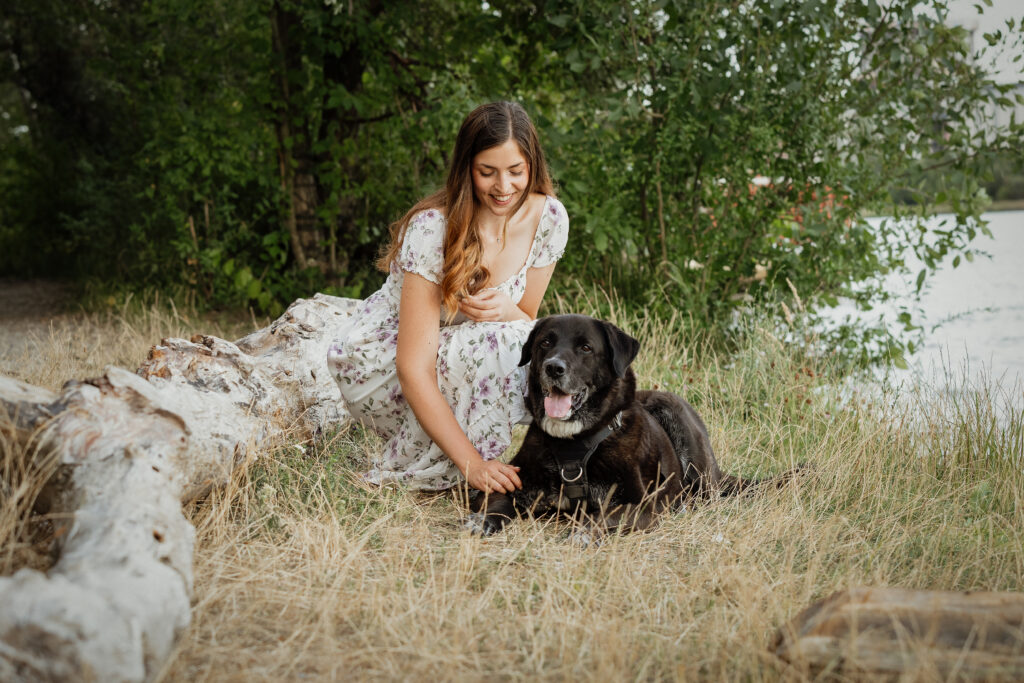 Schwarzer Hund liegt im Gras und seine Besitzerin beugt sich zu ihm runter.