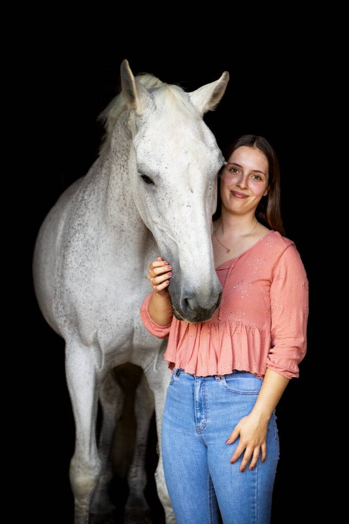 Weißes Pferd mit junger Besitzerin vor schwarzem Hintergrund im Stall.